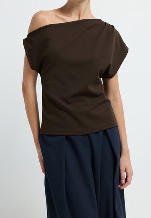 Femme portant un haut marron foncé dévoilant les épaules et un pantalon bleu marine ample sur un fond clair uni.