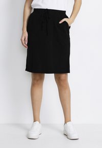 Jupe noire longueur genoux avec une taille à cordon de serrage, poches latérales et une texture lisse. Associée à des baskets blanches sur un fond uni.