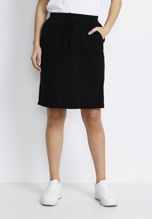 Jupe noire longueur genoux avec une taille à cordon de serrage, poches latérales et une texture lisse. Associée à des baskets blanches sur un fond uni.