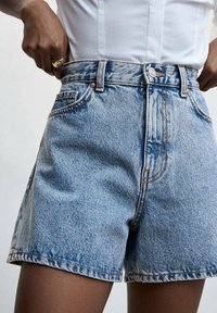 Shorts de mezclilla en azul claro con tiro alto, que cuentan con bolsillos frontales, herrajes de botón de cobre y detalles de costura visibles.