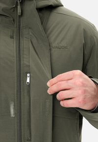 Giacca verde oliva, tessuto morbido, chiusura frontale con zip, cappuccio regolabile e chiusure in velcro. Presenta un ricamo del logo sul petto.