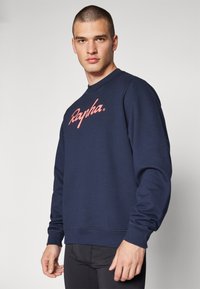 Sudadera azul marino hecha de tela suave, con puños y dobladillo acanalados; presenta un gran logo rosa "Rapha" en el pecho.