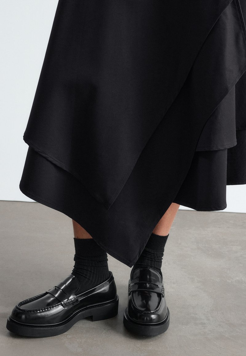 Mocassins en cuir noir à semelles épaisses portés avec des chaussettes noires et une jupe noire superposée sur un sol en béton gris.