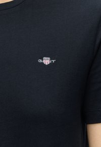 T-shirt en coton bleu marine avec un petit logo GANT brodé représentant un écusson blanc, rouge et bleu sur la poitrine gauche.
