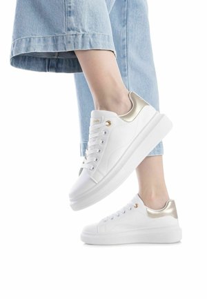 Zapatillas blancas con un acabado suave, detalles dorados en el talón y suelas planas. Los jeans del modelo son de color azul claro, recortados a la altura del tobillo.