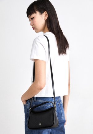 Jonge vrouw in wit T-shirt en blauwe spijkerbroek draagt een zwarte Calvin Klein schoudertas met geribbelde structuur aan haar rechterzijde.