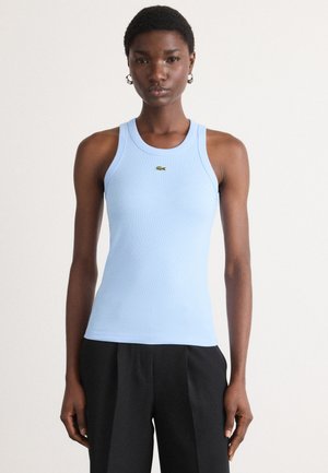 Débardeur côtelé bleu clair avec un col rond et des bretelles larges, arborant un petit logo vert Lacoste sur la poitrine. Porté avec un pantalon sombre.