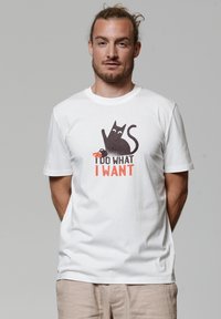 Witte katoenen T-shirt met een afbeelding van een zwarte kat met een krab, en de tekst "IK DOE WAT IK WIL" in oranje onder het ontwerp.