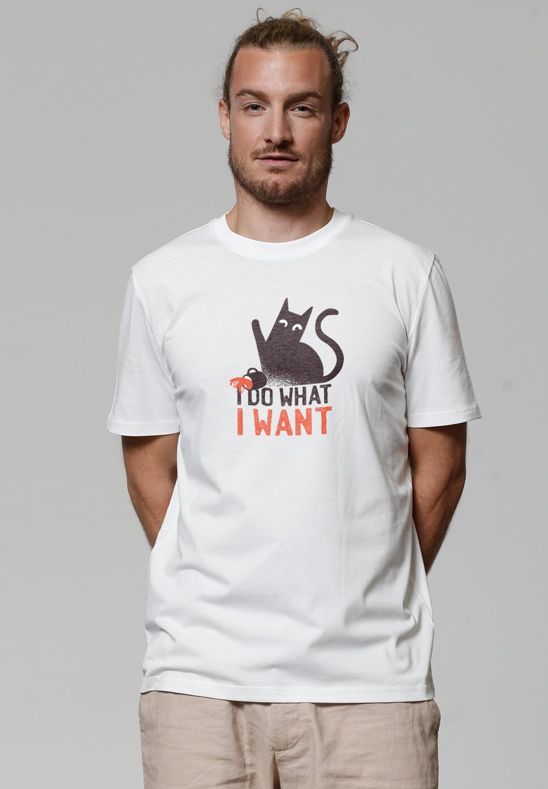 Witte katoenen T-shirt met een afbeelding van een zwarte kat met een krab, en de tekst "IK DOE WAT IK WIL" in oranje onder het ontwerp.
