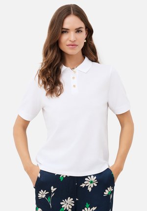 Femme aux longs cheveux bruns portant un polo blanc à manches courtes et un pantalon bleu marine à imprimé floral blanc, les mains dans les poches.
