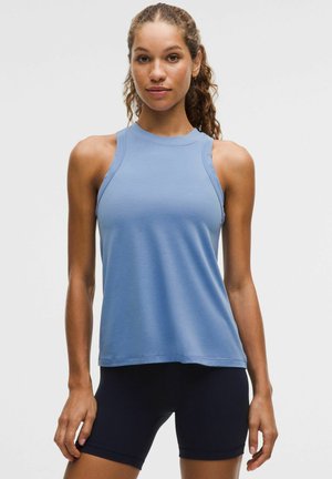 Kobieta z kręconymi włosami, ubrana w bezrękawnik w jasnoniebieskim kolorze oraz czarne dopasowane szorty, stojąca na jednolitym szarym tle.