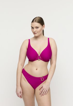 Vrouw met een lichte huid en haar in een staart, draagt een magenta bikini met gouden gespdetails, staand tegen een eenvoudige lichte achtergrond.
