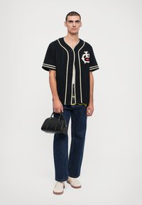 Fekete baseball póló fehér csíkokkal, emblémával, kék bővített nadrággal és bézs sportcipővel párosítva. A modellen egy fekete kézitáska van.