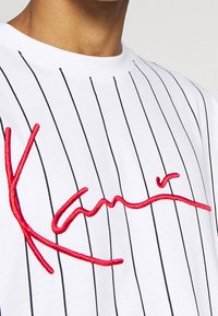 Bílý bavlněný tričko s tenkými černými vertikálními pruhy, se velkým červeným vyšitým nápisem uprostřed, který připomíná podpis.