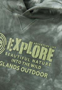 Tela texturizada verde con un estampado desvanecido que dice "Explora Aventura Vida Bella Naturaleza En la Selva Islas al Aire Libre."