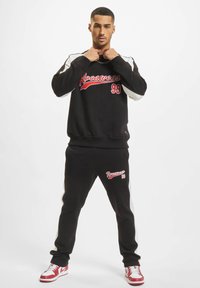 Juodas džemperis ir atitinkantys džinsai, su raudonais užrašais 'Rocawear 99'. Baltos detalės rankovėse, kartu su raudonai-baltomis sportinėmis bateliais.