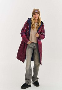 Długa pikowana kurtka w kolorze bordowym z kwiatowym wzorem, niebieska podszewka, noszona na beżowym swetrze z grubym splotem i jasnoszarych dzwonach jeansowych.