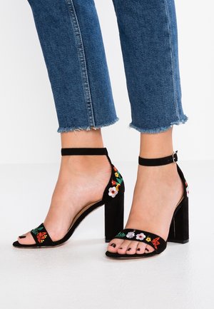 Sandales à talons en suède noir avec un bout ouvert, ornées d'une broderie florale colorée sur le côté et d'une sangle à boucle à la cheville.