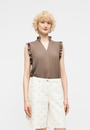 Kobieta o krótkich, kręconych blond włosach, ubrana w brązową, bezrękawnikową bluzkę z falbankami oraz białe, wzorzyste szorty do kolan na białym tle.