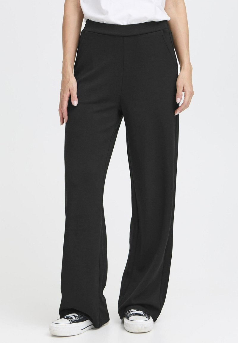 Pantalons larges noirs en tissu doux, avec une taille haute et des poches latérales, associés à des baskets blanches.