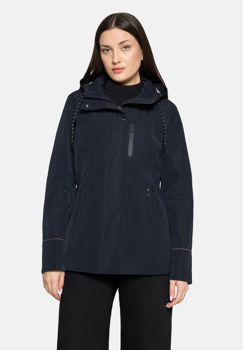 Veste imperméable marine avec capuche, fermeture éclair frontale, poches latérales et cordon de serrage ajustable. Présente un passepoil gris le long des coutures.