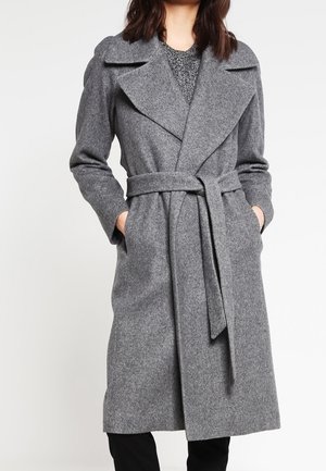 Vrouw draagt een lange grijze wollen jas met riem, brede revers en handen in de zakken, gecombineerd met een zwarte broek.