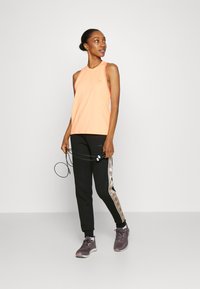 Haut de sport sans manches orange avec un bord contrasté, associé à un jogging noir avec une rayure décorative sur le côté et des baskets grises.