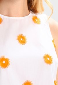 Gros plan sur un haut blanc sans manches avec une broderie florale circulaire orange vif sur la poitrine et la zone des épaules.