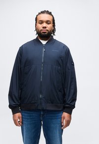 Άνδρας με πλεξούδες φοράει ένα μπλε σκούρο μπουφάν Hilfiger με φερμουάρ και μπλε τζιν, στεκόμενος μπροστά σε ένα απλό ανοιχτό φόντο.