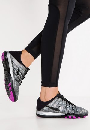 Person iført sorte leggings viser sølv- og sortfarvede Nike Free løbesko med pinksåler, den ene fod let løftet.