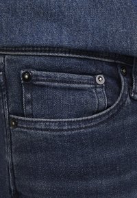 Close-up van een donkerblauwe denim spijkerbroekzak met zichtbare stiksels, klinknagels en gestructureerde stof.