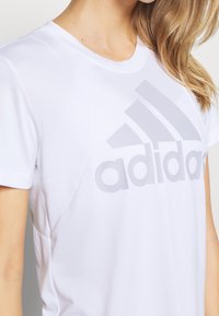 Camiseta atlética blanca de manga corta, con un gran logo de Adidas en gris claro en el frente y una textura de tela suave y ligera.