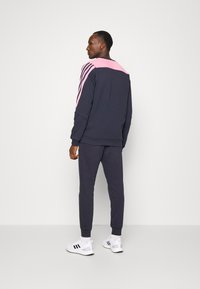 Sudadera azul marino con hombros rosas y tres rayas rosas; combinada con joggers azules marinos a juego y zapatillas blancas. Textura suave, diseño casual.