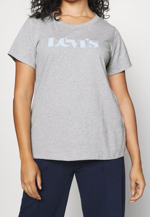 Kobieta ubrana w jasnoszarą koszulkę Levi's i ciemnoniebieskie spodnie, stojąca z ramionami swobodnie opuszczonymi wzdłuż ciała na jednolitym tle.