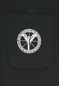 Schwarzer Stoff mit einem zentrierten weißen Logo, das eine geflügelte Figur in einem kreisförmigen Design zeigt, detailliert mit geometrischen Mustern am Rand.