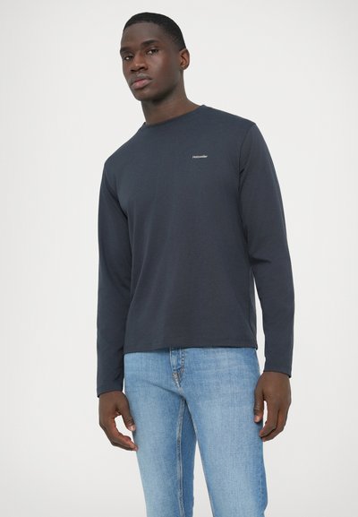 Camisola azul-marinho de manga comprida com decote redondo e pequeno logótipo, feita de algodão. Combinada com jeans azul claro, modelo posa contra um fundo liso.