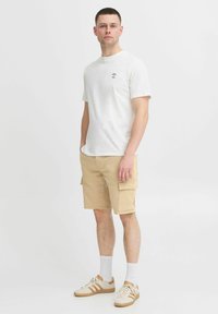 Camiseta blanca de algodón con un pequeño logo negro, pantalones cortos de cargo color beige con bolsillos laterales, calcetines blancos y zapatillas beige con detalles marrones.