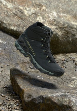 Botte de randonnée La Sportiva noire et grise avec Gore-Tex, affichée en suspension au-dessus d'un terrain rocheux.