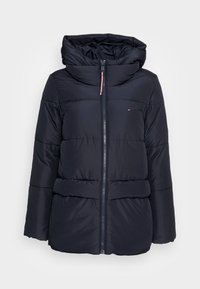 Marineblå pufferjakke med høj krave, lynlås-lukning og vandret syninger. Har en hætte med faux fur-foring og indvendig snøre.