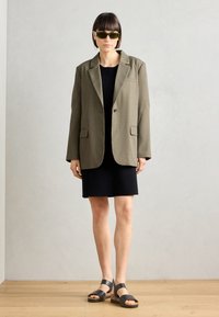Olive green oversized blazer with structured shoulders, black fitted dress, and black sandals with cork details. Smooth texture, minimal design.