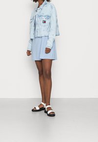 Ljusblå denimjacka i kort modell med fransad fåll, randig ljusblå kort klänning och vita sandaler med svarta sulor och ankelremmar.