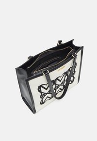 Stor tygväska med en krämfärgad tygkropp, svart broderad mönster, svarta konstläderdetaljer, guldfärgade beslag och dubbla handtag.