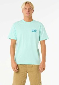 Camiseta de manga corta de color azul claro, hecha de algodón, con un pequeño gráfico y texto en el pecho izquierdo. Llevada con pantalones cortos color caqui.