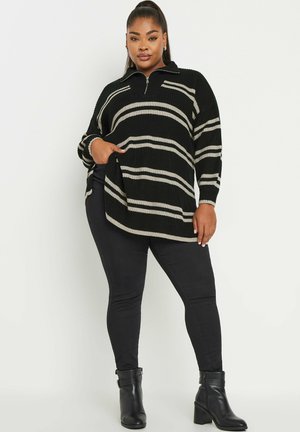 Suéter de punto rayado en negro y beige con cuello con cremallera, que presenta un corte relajado y bolsillos laterales, combinado con jeans ajustados negros y botas de tobillo.