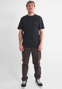 T-shirt noir avec un petit logo sur la poitrine, associé à un pantalon marron et des baskets noir et blanc, se tenant devant un fond blanc.