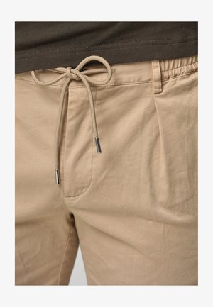 Beige bomuldsbukser med snoreliv, der har metalender. Stoffet er glat med subtile krøller og sidelommer.