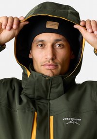 Grön vattentät jacka med gula detaljer, hög krage och logotyp. Svart stickad mössa som bärs under huvan, vilket visar på en snug passform.