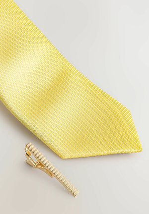 Gul slips med en texturerad väv, tillsammans med en guldtonad slipsklämma med ett mönstrat yttre och en smal, rektangulär form.