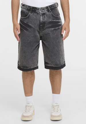 Mand iført sorte vaskede denimshorts, hvide sokker og beige-hvide sneakers stående mod en ensfarvet baggrund.