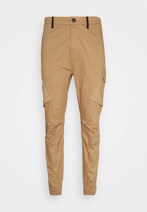 Tan slim-fit cargo pants with button and zip closure, flap pockets on thighs, elastic cuffs, and black belt loops.